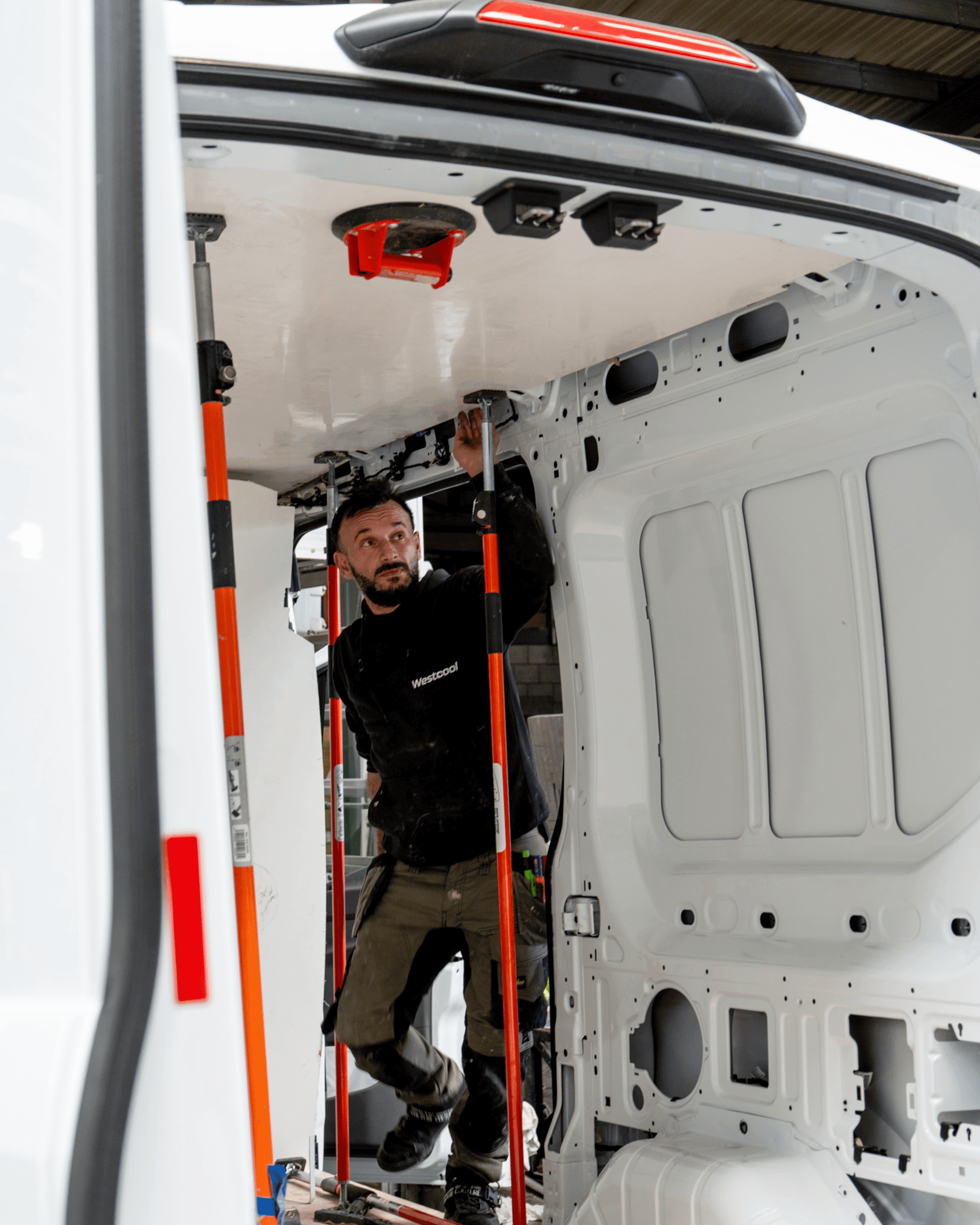 A technician clamping an insulation panel against the roof of a white van under conversion, using orange props