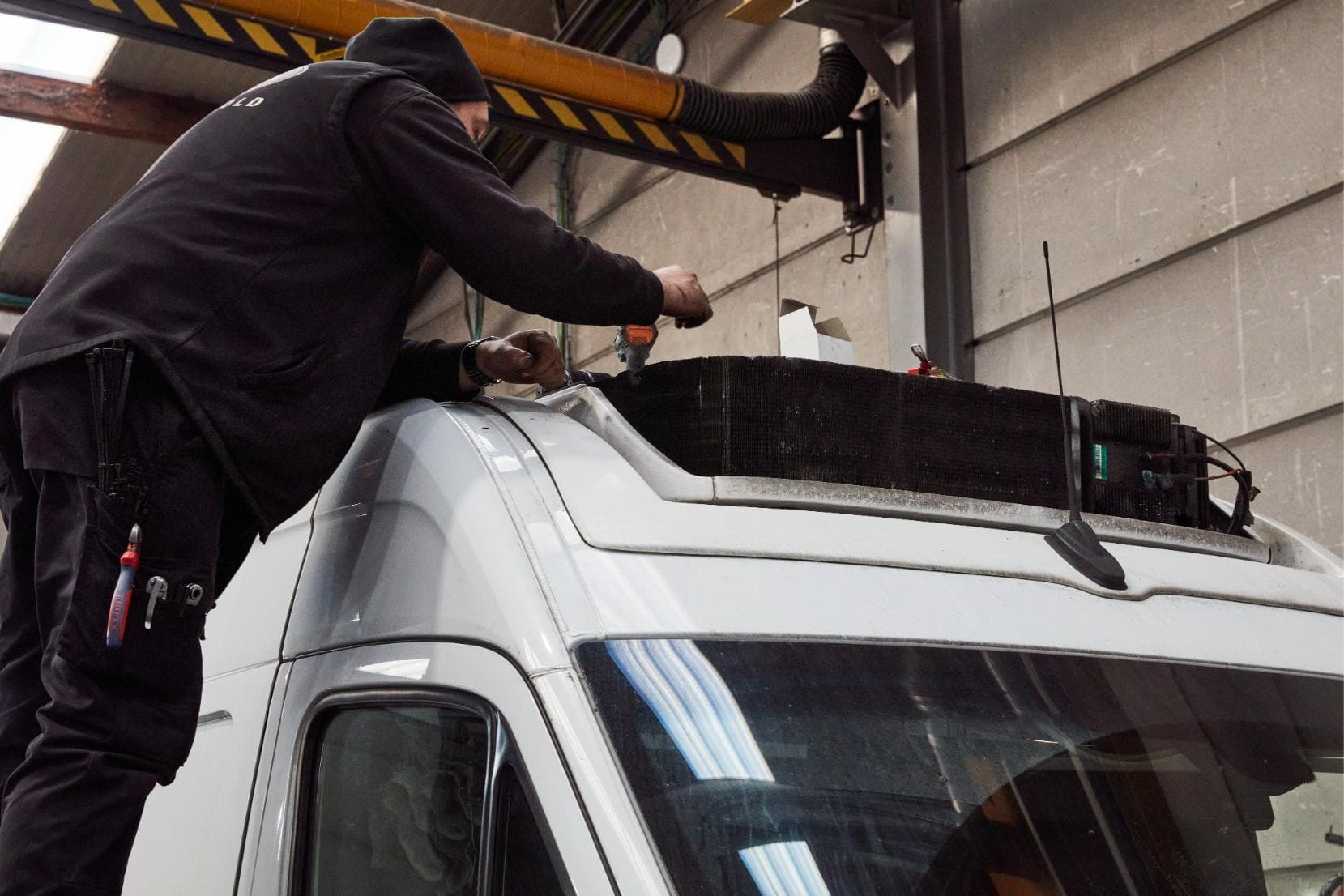 A coachbuilder on a stepladder cutting open the roof of a white panel van for the refrigeration unit, the evaporator block already sitting on the roof beside him