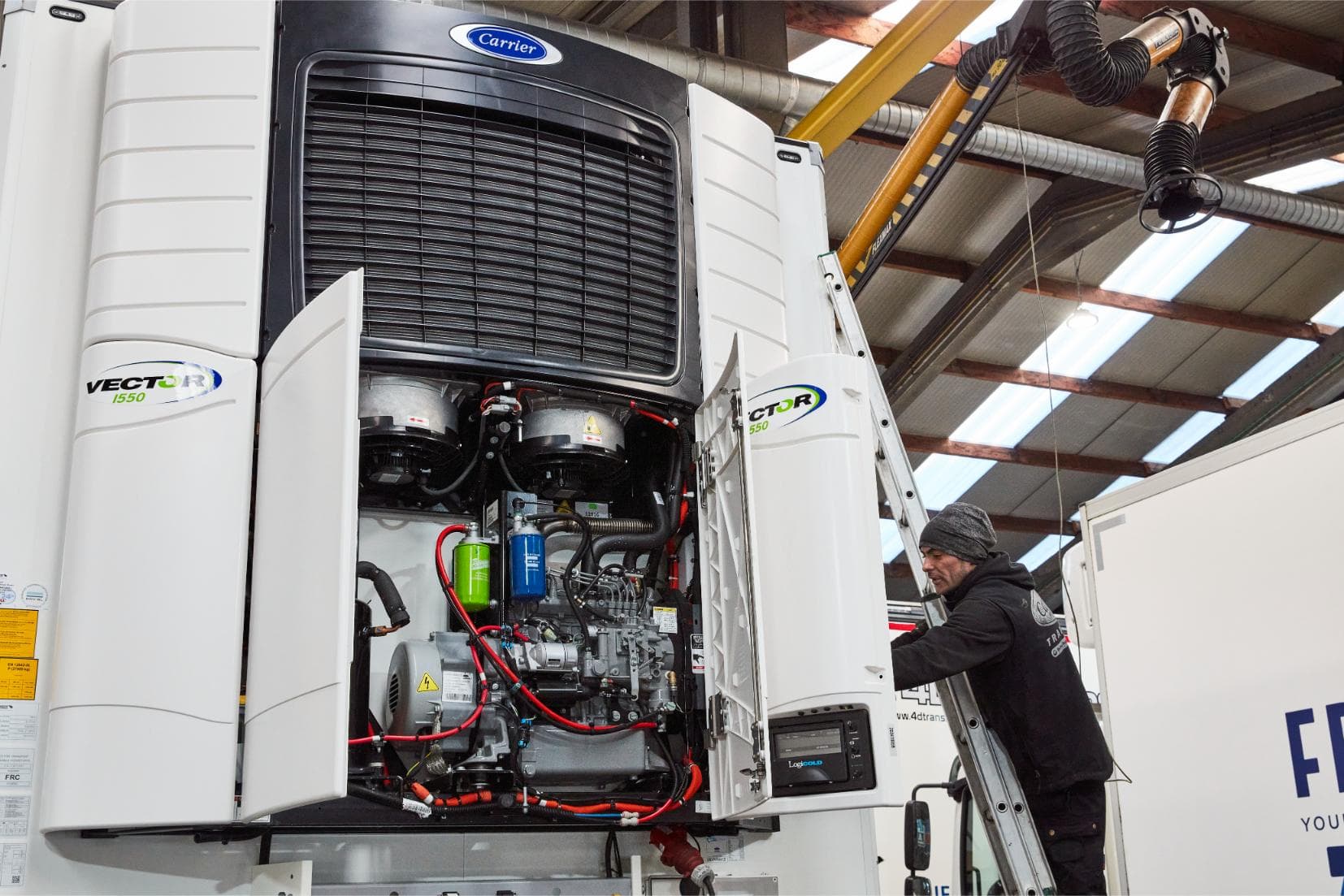 A Carrier technician on a ladder beside an opened refrigeration unit on the roof of a truck in the workshop