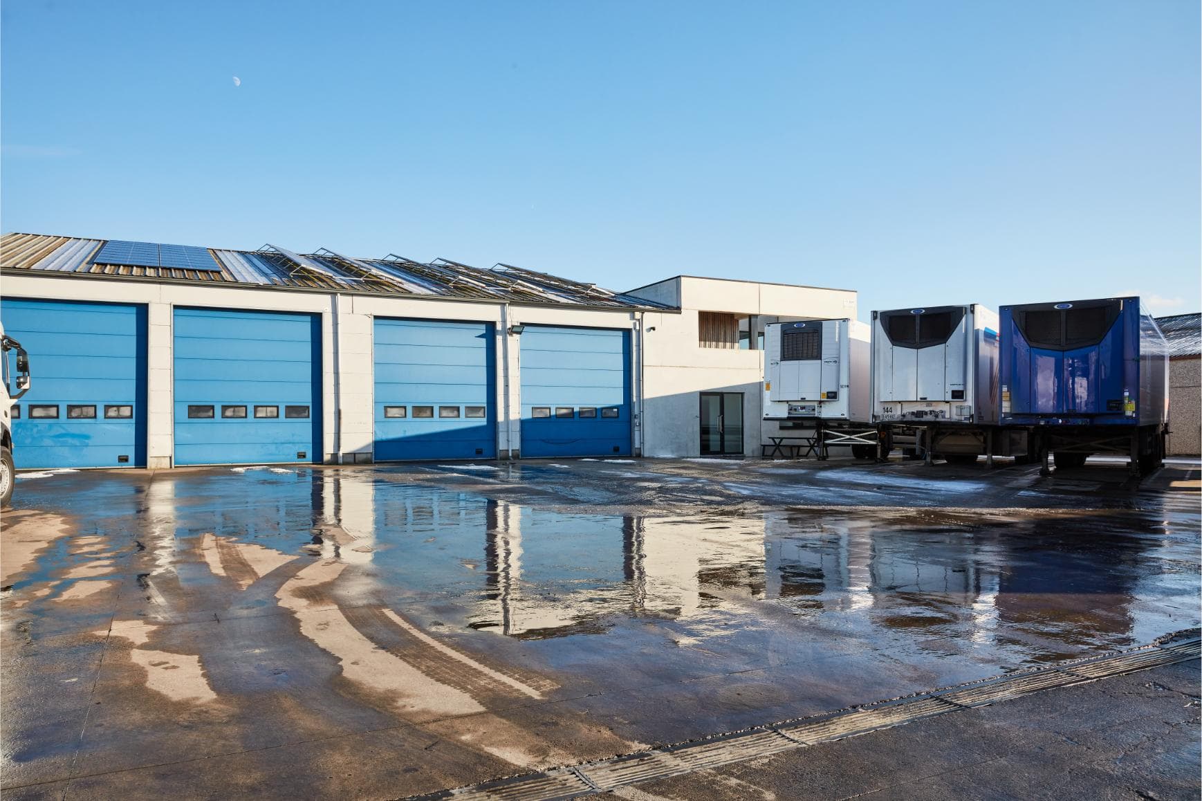 The Hooglede site from the yard: four blue roller doors, solar panels on the roof and refrigerated trailers on the yard